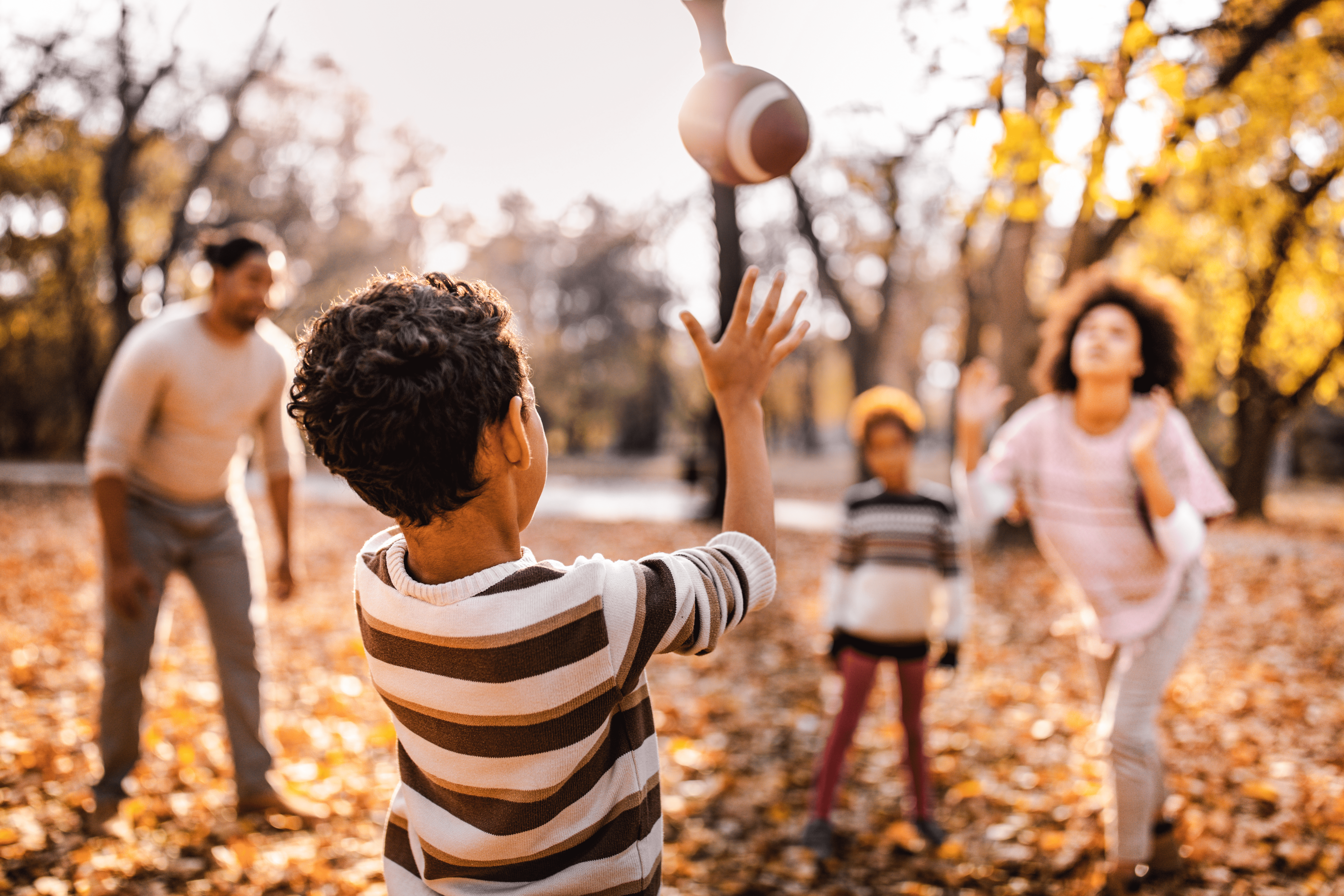 Un grupo de cuatro personas (un hombre, una mujer y dos niños) lanzan un balón de fútbol en un paisaje otoñal al aire libre.  