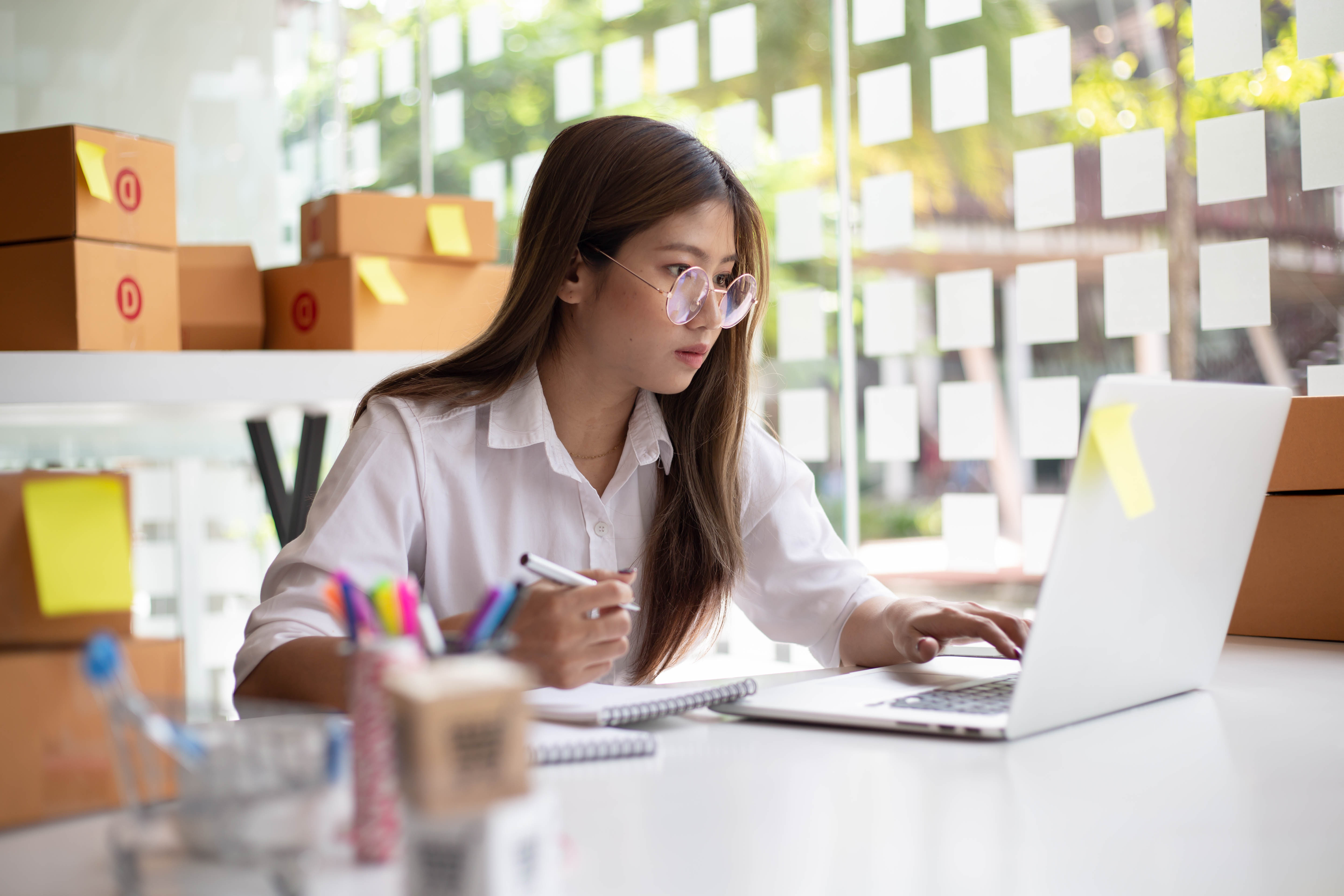 Asian woman sitting at a desk looking at a laptop with cardboard packing boxes behind her.