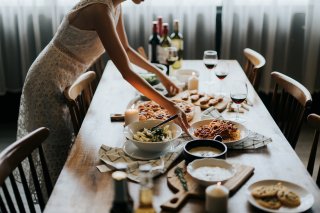 A woman in a summery white dress sets dishes of pasta, soup, pizza, and appetizers on a wooden table. On the table one can see wine glasses and bottles with red wine, as well as lit wax candles, and plaid napkins, and bowls of soup.   