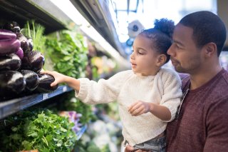 A toddler held in her father's arms reaches for a shiny eggplant in the produce section of a market, amidst an array of leafy green vegetables, as the dad looks on encouragingly. 