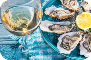 A plate of raw oysters with a slice of lemona, and a glass of white wine are served on a blue dish with a blue and white checkered napkin.   