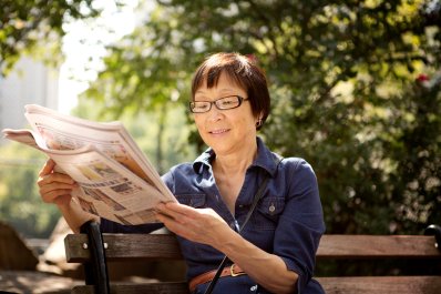 A smiling woman sitting on a bench reading a newspaper