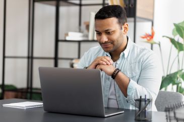 A smiling man sitting in front of an open laptop at a desk” 