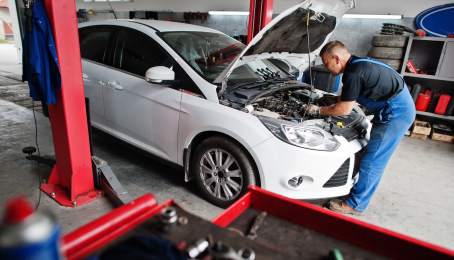 A man working under the hood of a white car in an autobody shop.