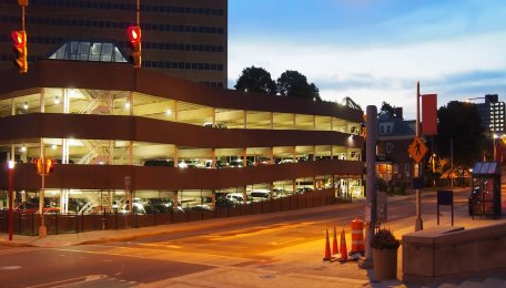 A parking garage that is lit up on the inside, with a city street and the sunset in the background.