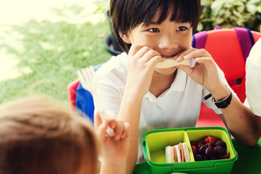 a young Asian boy eating a sandwich from a green lunch box